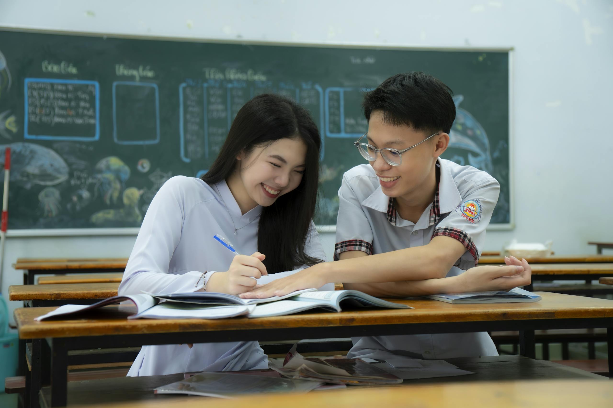 Two high school students study together in a classroom, smiling and engaged.